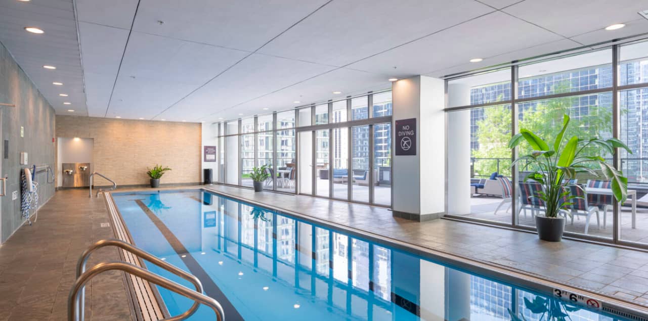 Indoor lap pool with blue water, metal railings, large windows showing downtown Chicago buildings, potted plant, and recessed ceiling lights.