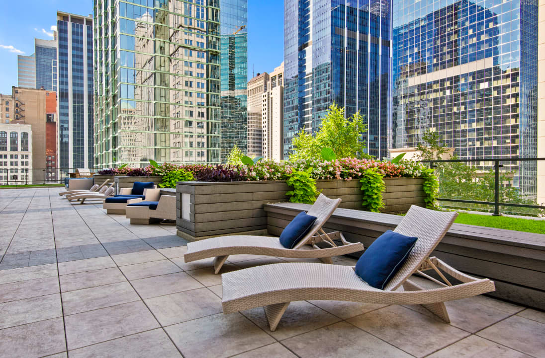 Rooftop terrace with woven lounge chairs, blue cushions, wooden planters with flowering plants, and Chicago skyline views.