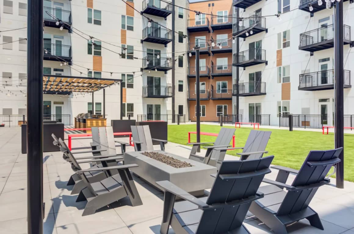 Courtyard with modern black and gray lounge chairs arranged around a linear fire pit, surrounded by multi-story apartment buildings with balconies and a bright green lawn.