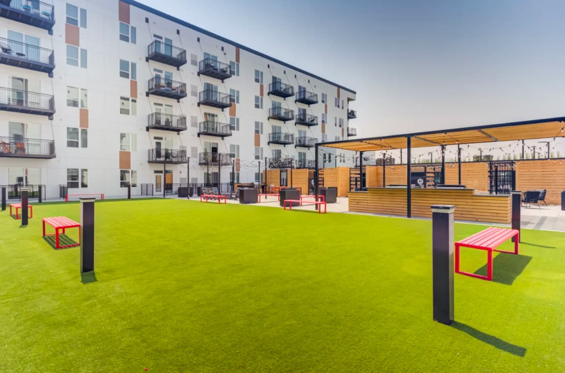 Outdoor courtyard with bright green lawn, red and pink benches, black metal posts, modern apartment building with balconies, and wooden pavilion structure.