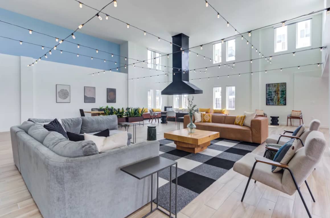 Spacious loft-style common room with gray and tan sectional sofas, black pendant hood, string lights, large windows, and modern furnishings arranged throughout.
