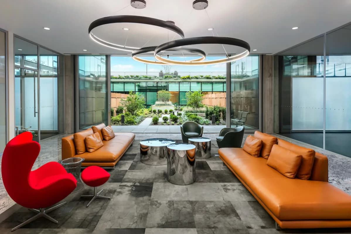 Contemporary lounge with orange leather sofas, red accent chair, metallic side table, circular black pendant lights, and expansive windows overlooking green courtyard.