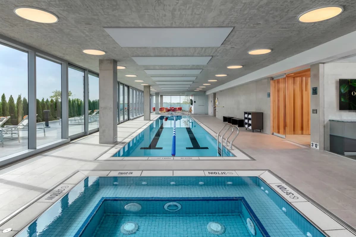 Indoor swimming facility with two pools, concrete ceiling with recessed lighting, large windows, and lounge seating area.