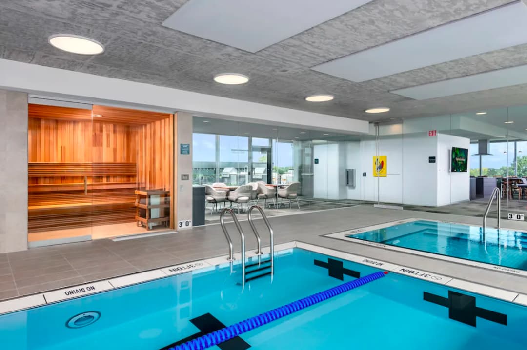 Indoor swimming facility with two lap pools separated by lane dividers, wooden sauna room on left, lounge seating in background, and recessed ceiling lights.