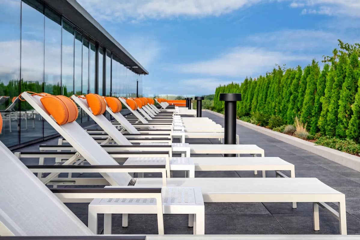 Rooftop terrace with white lounge chairs, orange cushions, glass railing, and evergreen trees under blue sky.