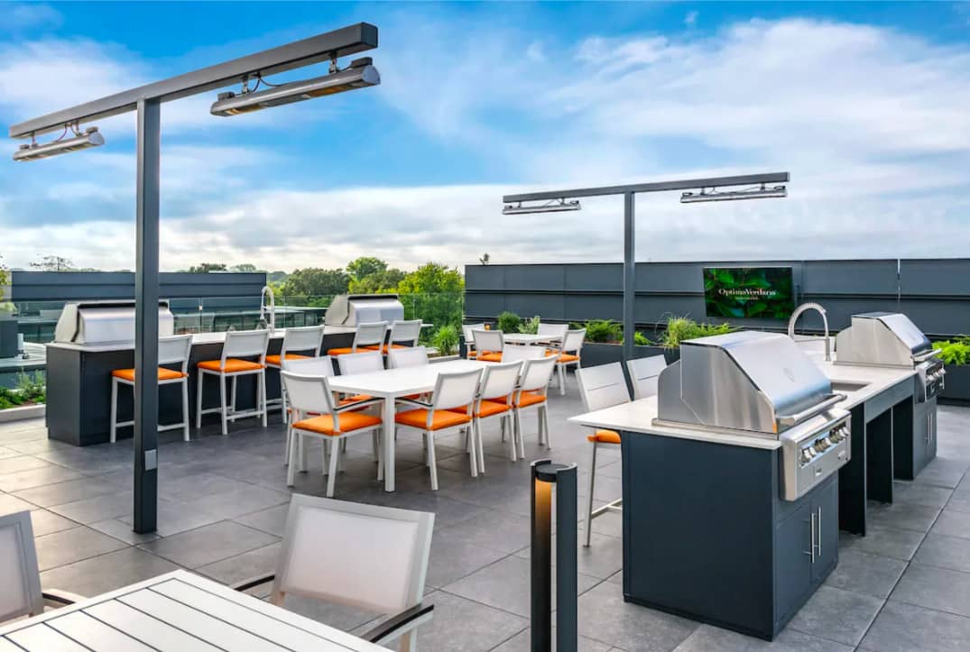Rooftop terrace with stainless steel grills, white dining tables with orange chairs, overhead heaters, and city views.