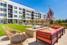 Outdoor courtyard with woven lounge chairs, fire pit, cornhole game, and modern apartment building in background under clear sky.