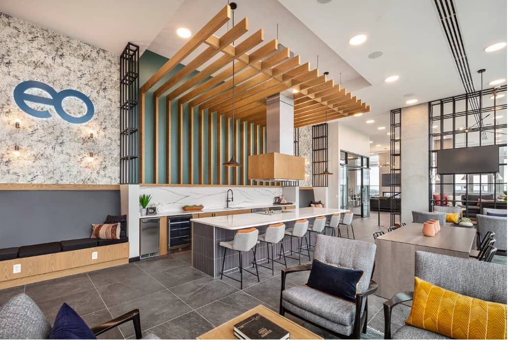 Open-concept common area with wood beam ceiling, marble accent wall, kitchen with white countertops, dining counter with bar seating, and gray upholstered lounge furniture.