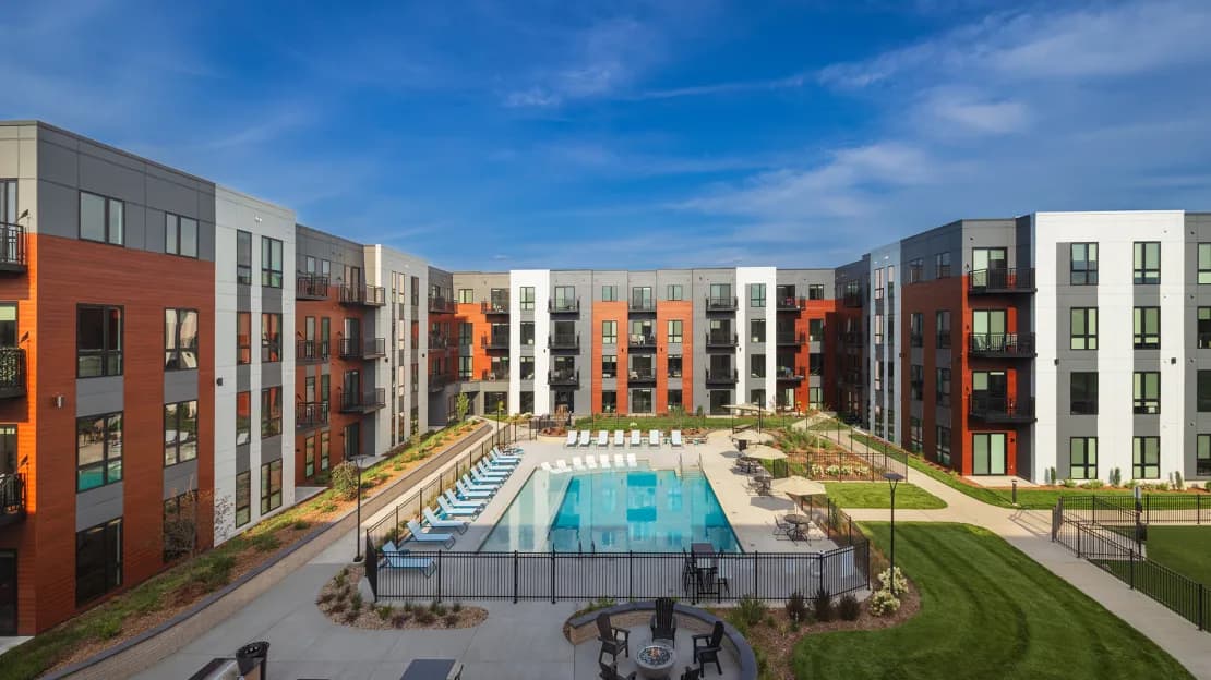 Aerial view of apartment complex courtyard with rectangular pool, lounge chairs, manicured lawn, and multi-story residential buildings with orange, gray, and white facades.