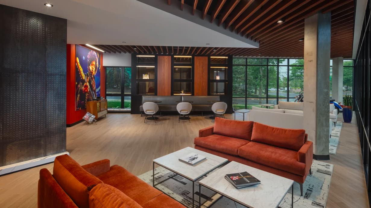 Open-concept lounge with orange sofas, white coffee tables, wood-slat ceiling, and floor-to-ceiling windows with views of landscaped grounds.