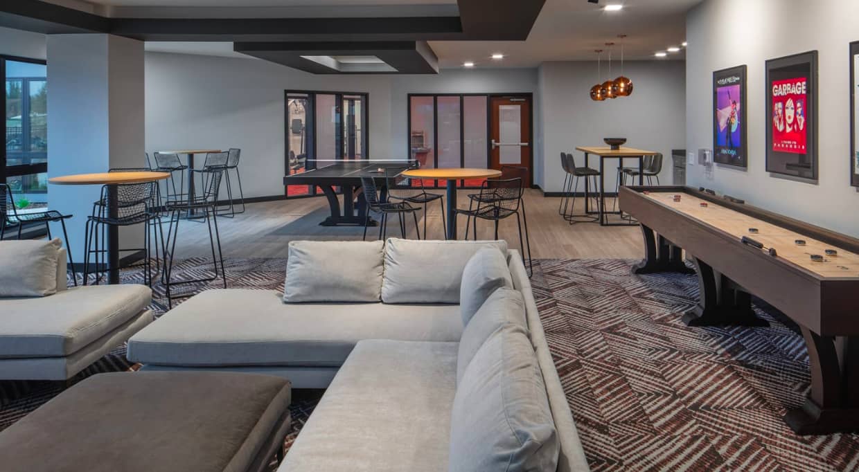 Open lounge area with light gray sectional sofa, pool table, shuffleboard, high-top bar tables with black chairs, and geometric patterned flooring.