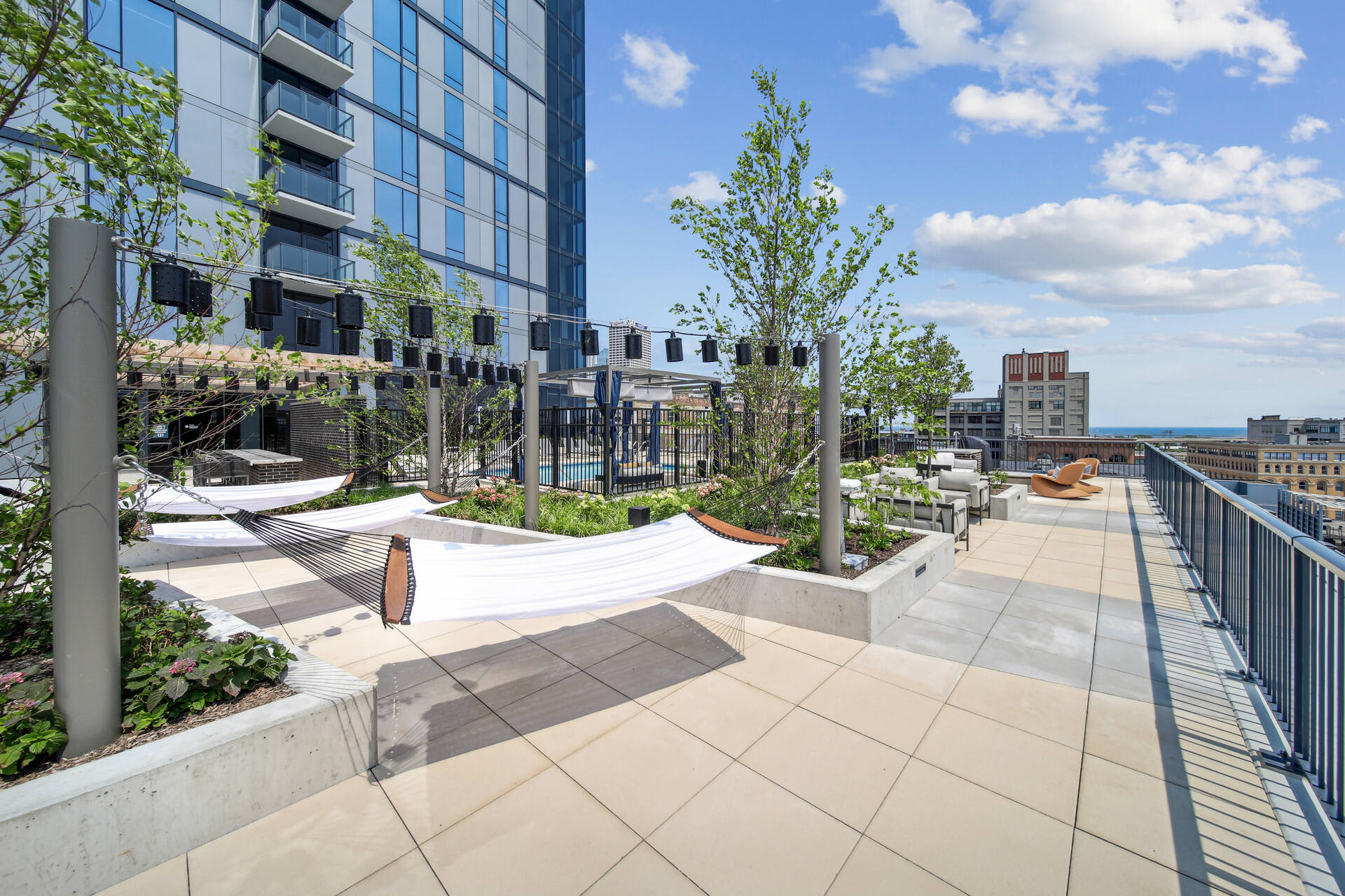 Rooftop terrace with white shade structures, planted trees, lounge seating, and metal railing overlooking Milwaukee skyline.