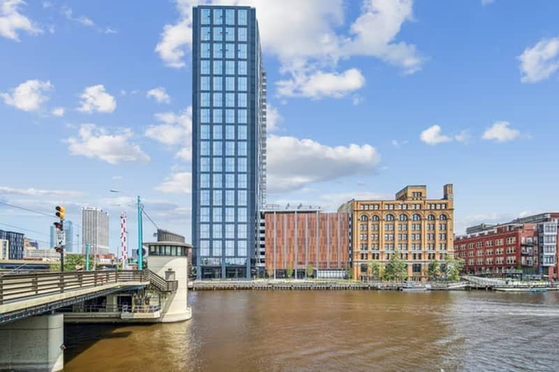 Tall modern glass tower on the Milwaukee River waterfront, flanked by historic brick buildings and a pedestrian bridge.
