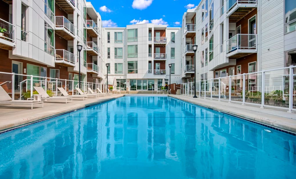 Resort-style swimming pool with clear blue water, white lounge chairs, and modern multi-story apartment buildings with balconies surrounding the courtyard.