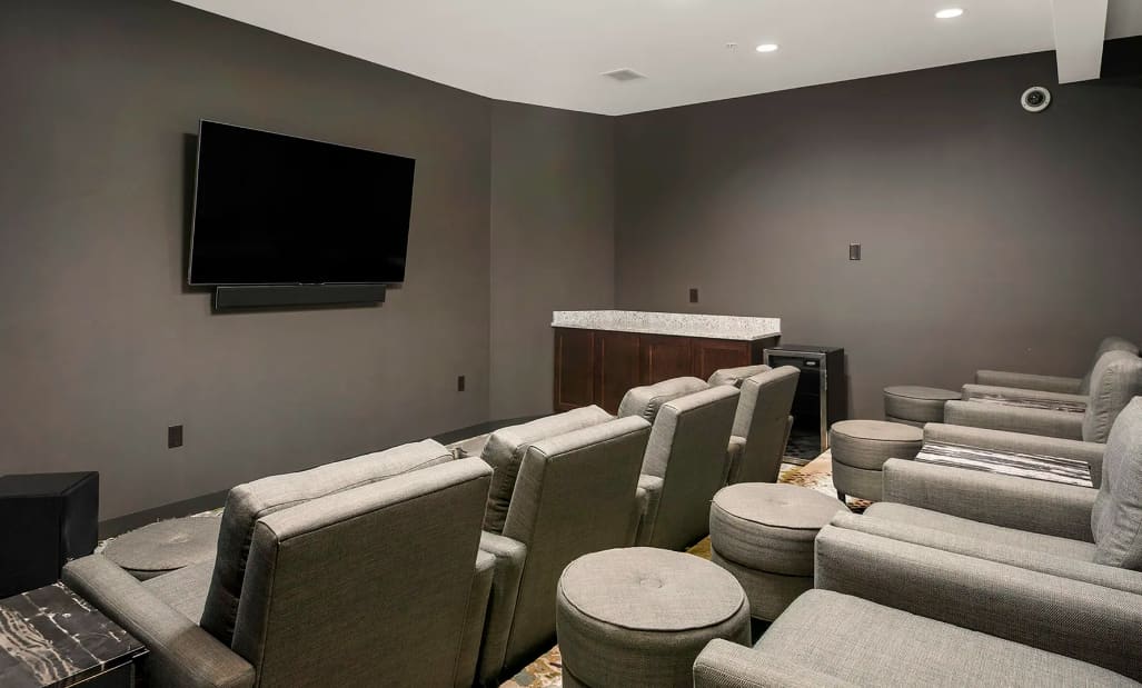 Home theater room with multiple rows of beige upholstered recliners, wall-mounted flat-screen TV, and dark accent walls with recessed lighting.