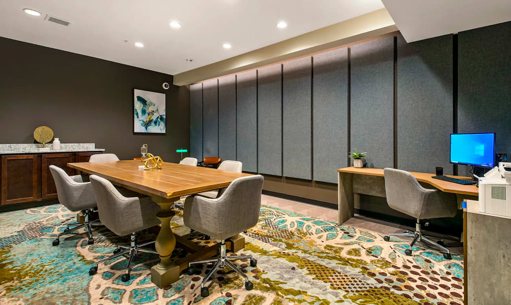 Conference room with wooden table, gray upholstered chairs, dark green walls, patterned rug, and built-in desk workspace with monitor.