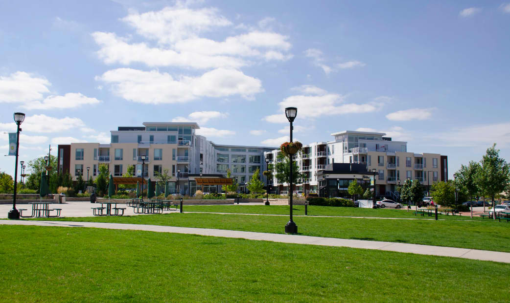 Multi-story residential buildings with mixed-color facades surrounding a large open green lawn, concrete pathways, and black street lamps under blue sky.