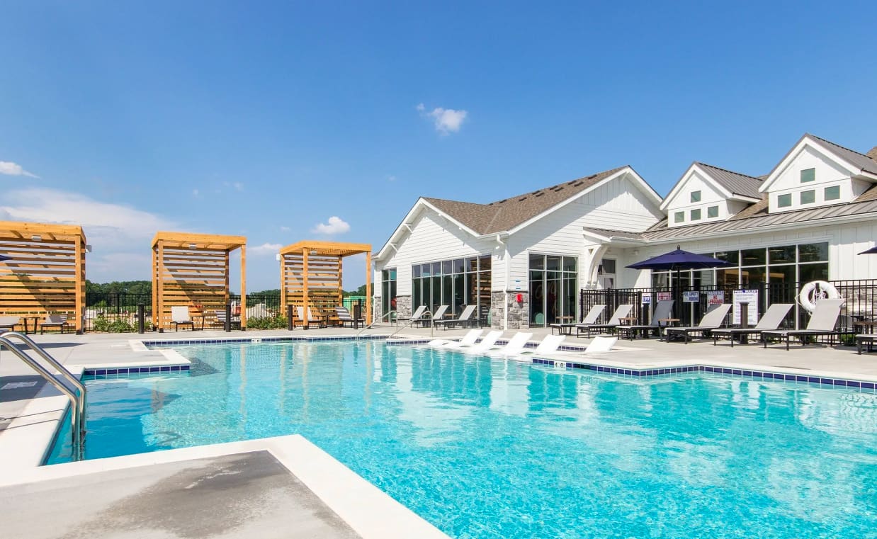 Resort-style swimming pool with clear blue water, white lounge chairs, wooden pergola structures, and white clubhouse building with large windows under blue sky.