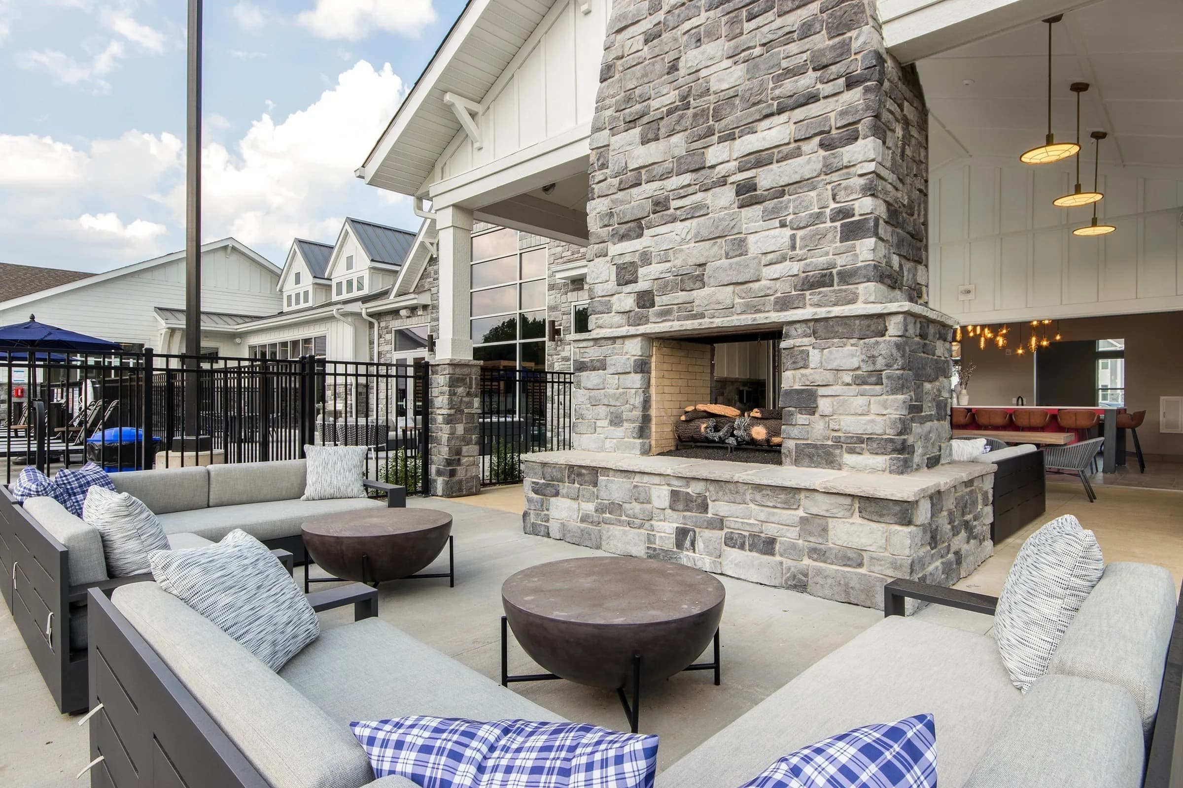 Outdoor patio with gray upholstered seating, stone fireplace with opening, two dark fire bowls, pendant lights, and view of residential buildings.