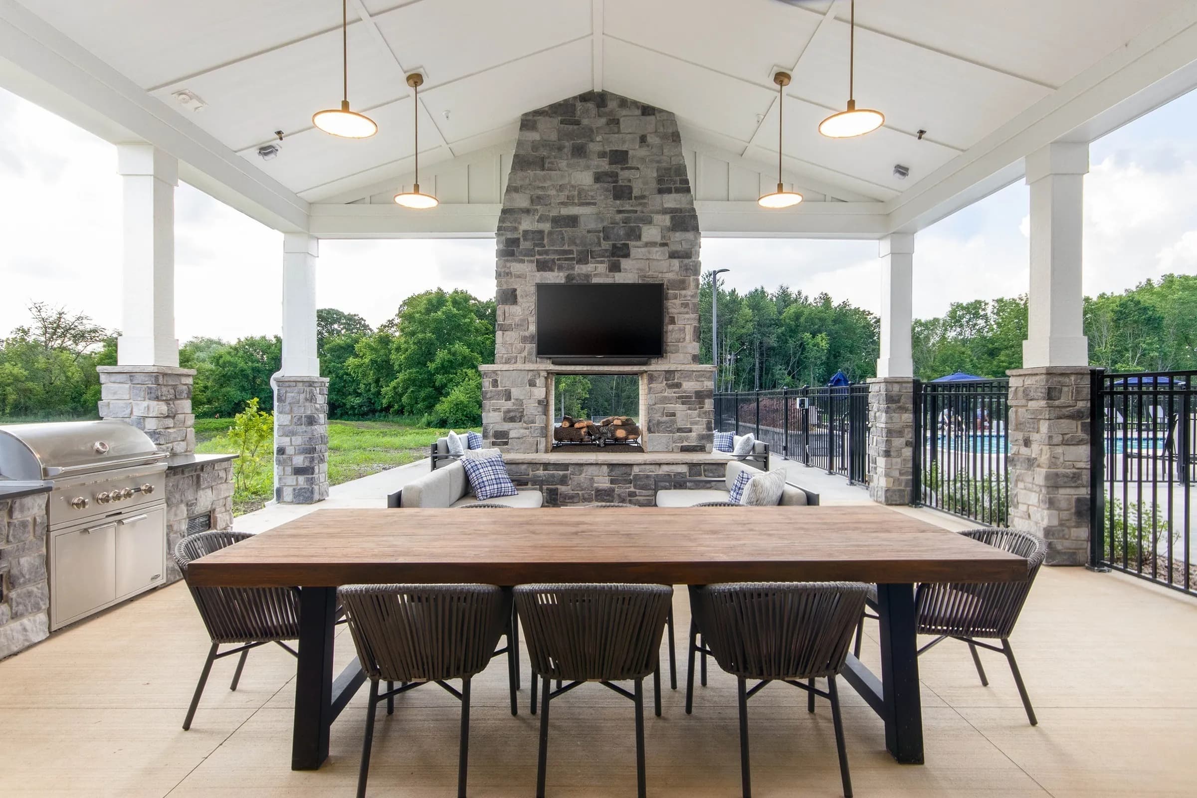 Covered pavilion with wooden dining table, black chairs, stone fireplace with TV, pendant lights, and views of trees and lawn.
