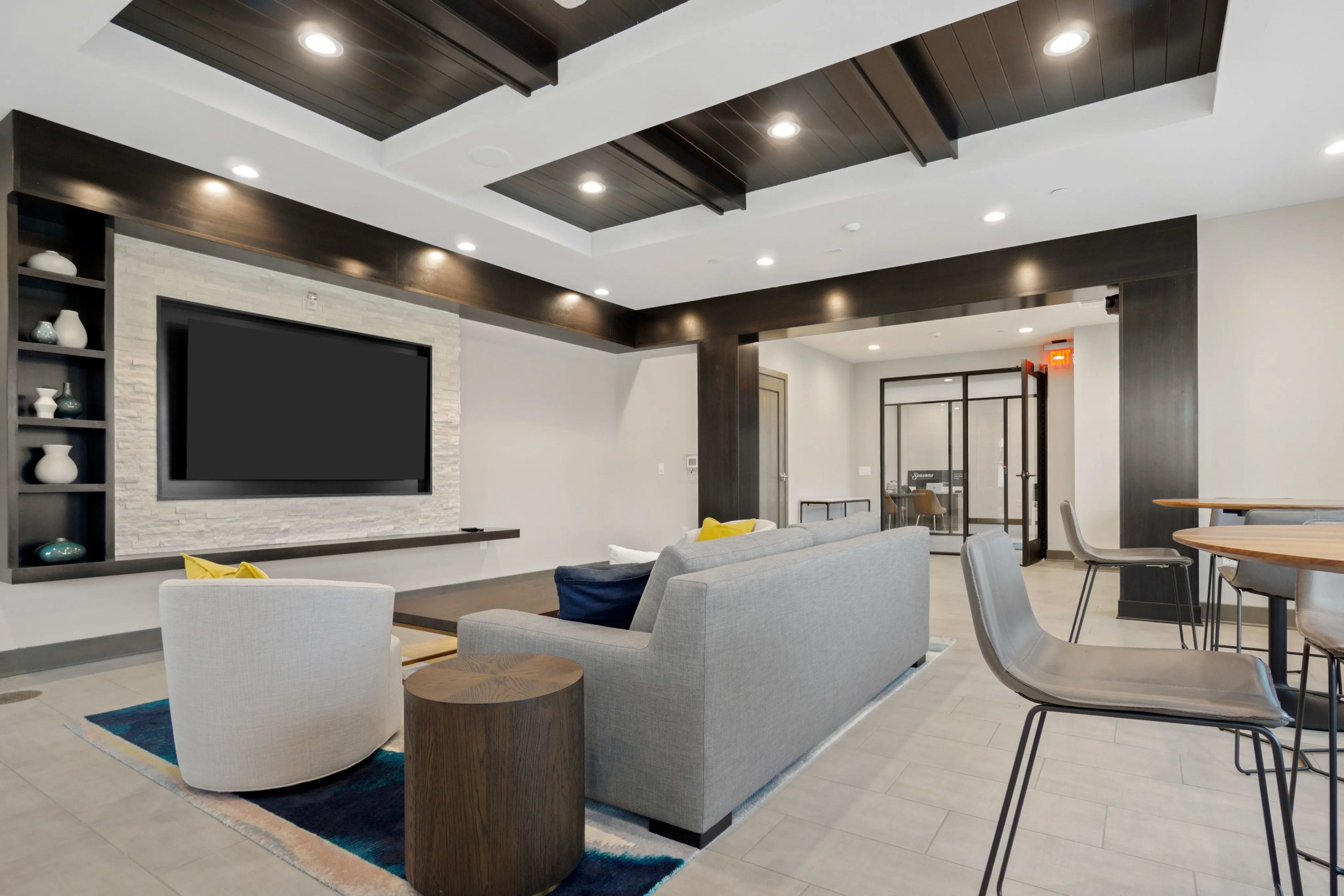 Media room with gray L-shaped sectional, large wall-mounted TV, dark wood ceiling beams, and adjacent dining area with wood table and chairs.
