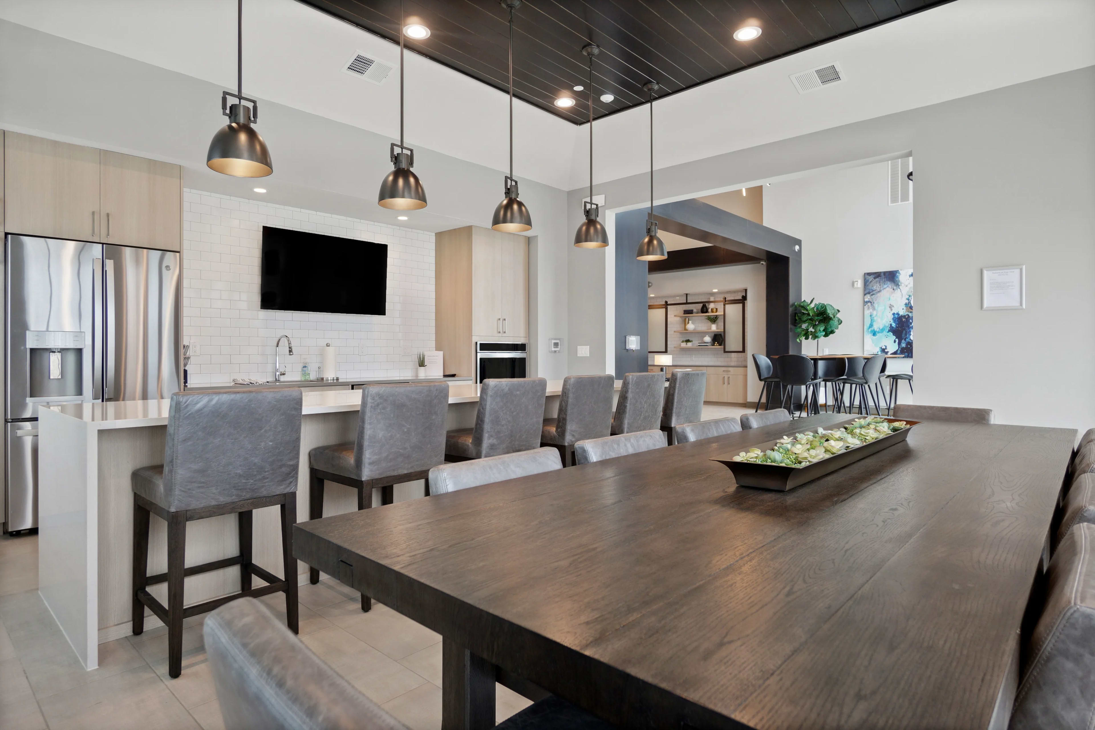 Open-concept dining and kitchen with large wood dining table, gray upholstered chairs, pendant lights, white subway tile backsplash, and stainless steel appliances.