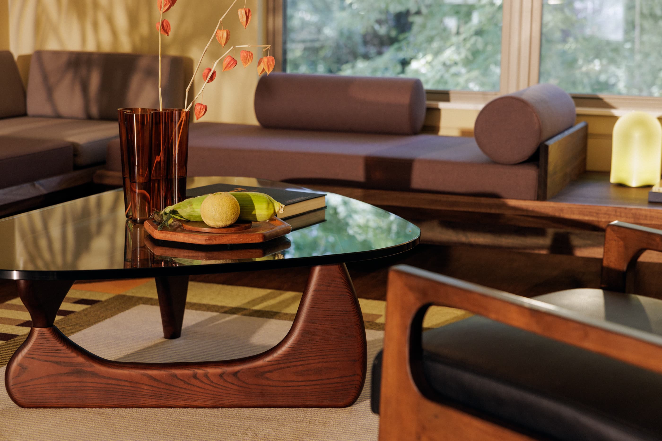 Living room with glass-top coffee table, brown leather sofa, wooden mid-century furniture, and large windows overlooking greenery.
