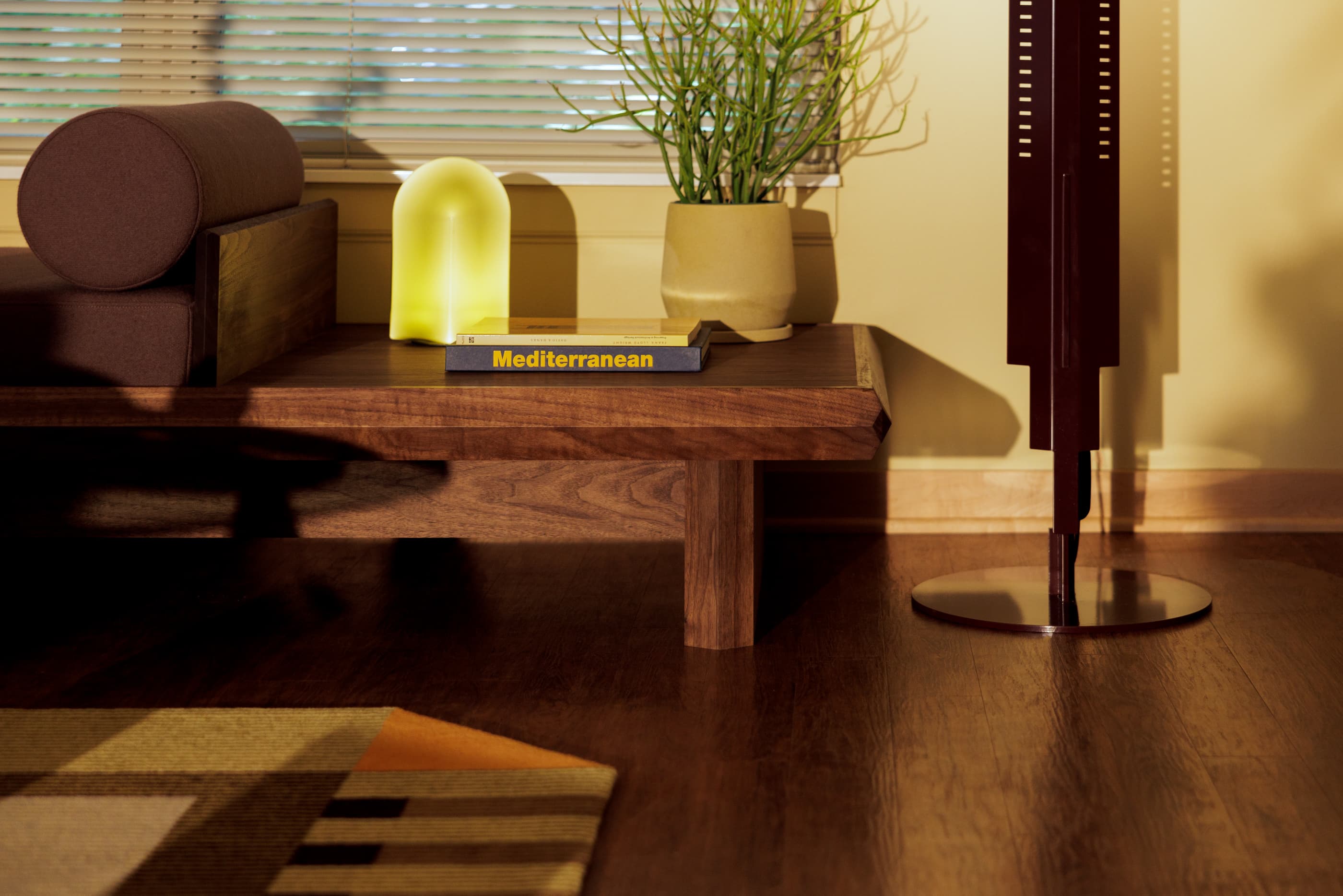 Living room with dark wood coffee table, brown accent pillows, glowing yellow lamp, potted plant, and wooden shutters with natural sunlight.
