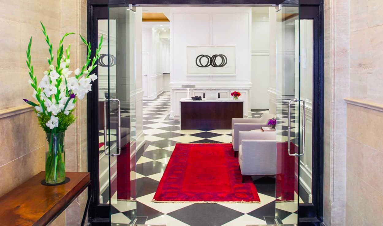 Lobby entrance with black glass doors, red geometric rug, white flowers in vase, dark reception desk, and diamond-patterned floor.