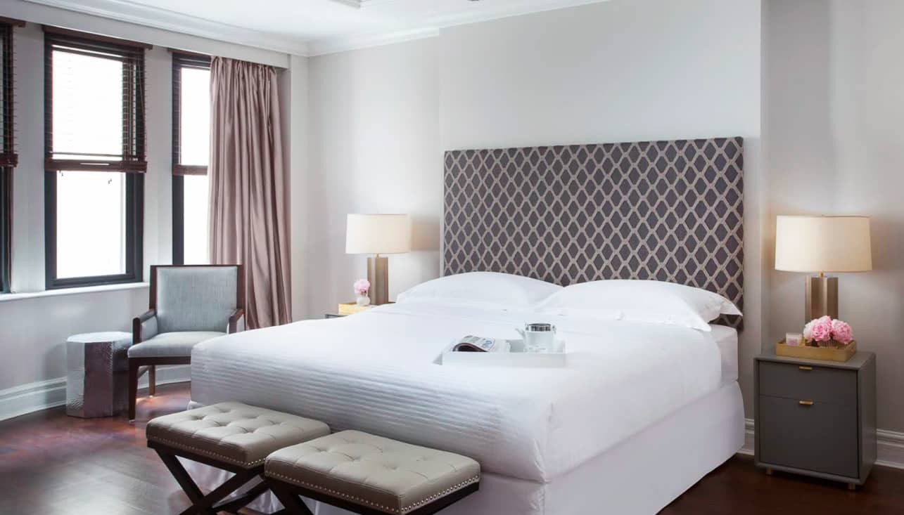 Bedroom with white bedding, gray patterned headboard, two brass table lamps on dark nightstands, large windows with taupe curtains, and upholstered bench at foot of bed.