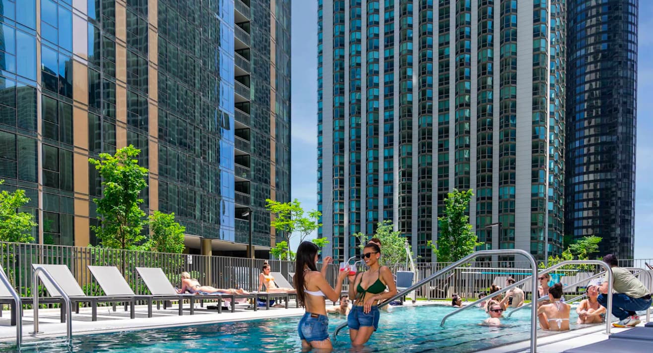 Rooftop pool with swimmers and lounge chairs, surrounded by tall residential buildings and green trees under blue sky.