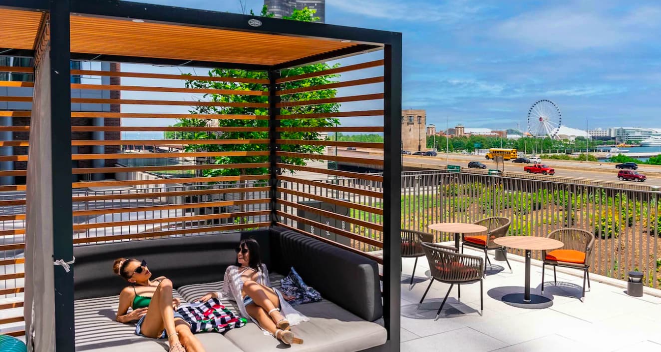 Two people relaxing on a cushioned daybed under a black pergola with wooden slats, overlooking Chicago's lakefront and skyline.