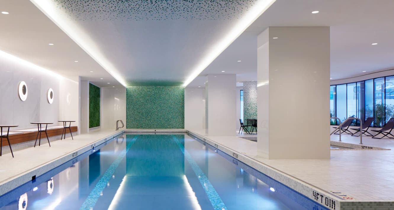 Indoor lap pool with turquoise water, green tile feature wall, recessed ceiling lights, and lounge seating area visible in background.