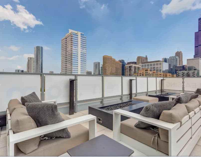 Rooftop terrace with built-in beige seating, fire pit, and Chicago skyline views behind glass railing.