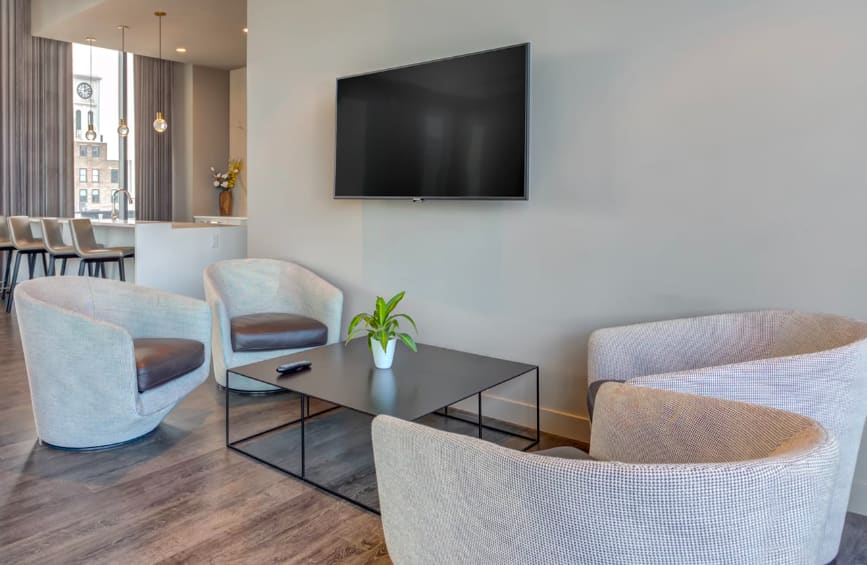 Living room with four cream upholstered chairs, black coffee table, wall-mounted TV, and large windows with city views.