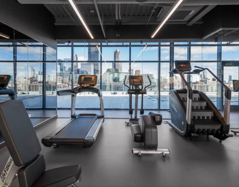 Modern gym with treadmill, elliptical, and weight equipment facing expansive windows with Chicago skyline view.