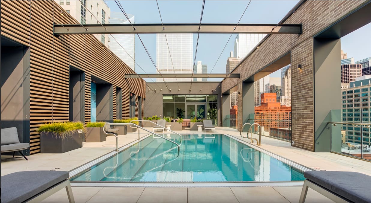 Rooftop lap pool with glass railings, lounge chairs, wooden slatted walls, and Chicago skyline views under glass ceiling.