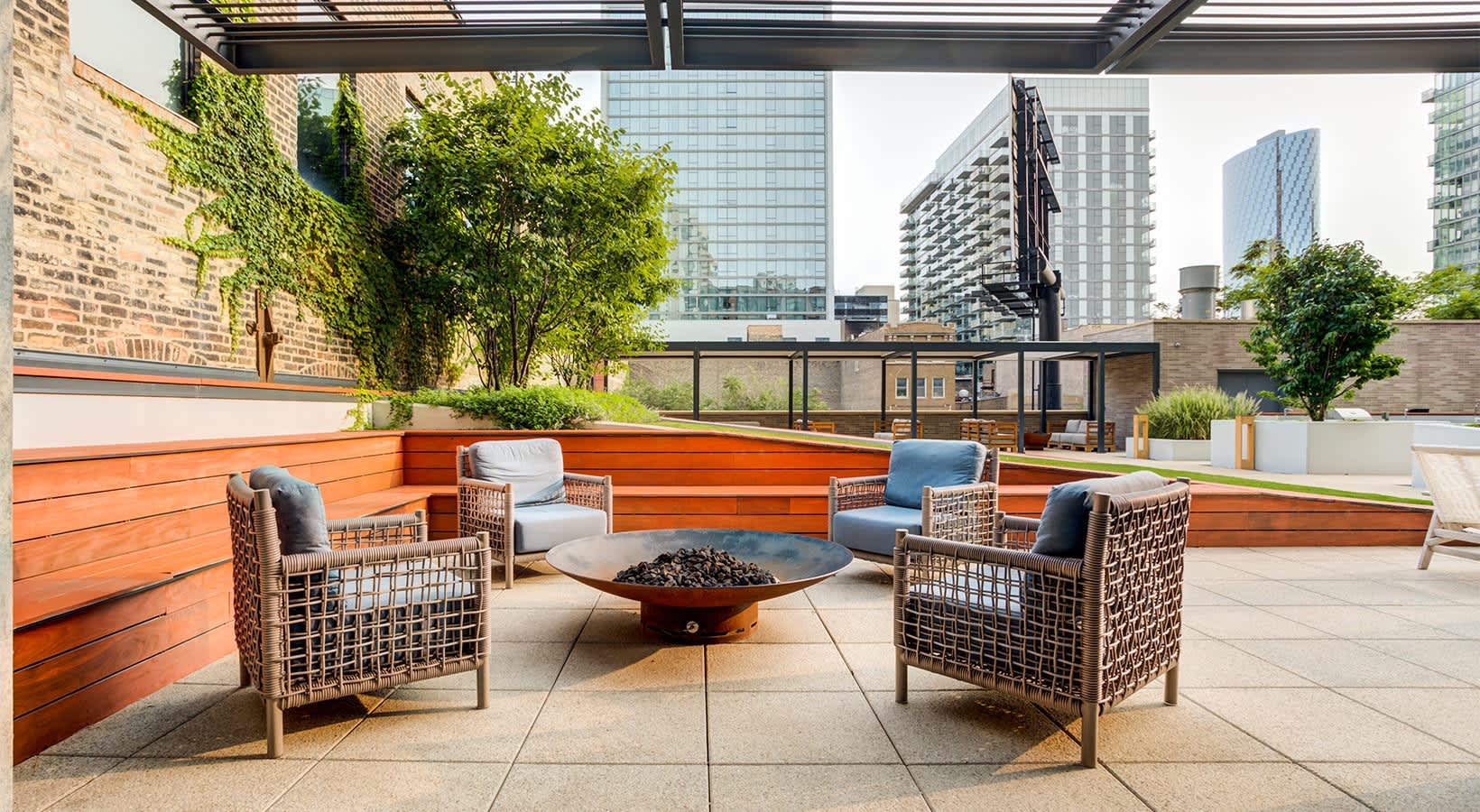 Rooftop terrace with woven lounge chairs, orange wooden benches, central fire pit, and downtown Chicago skyline backdrop.