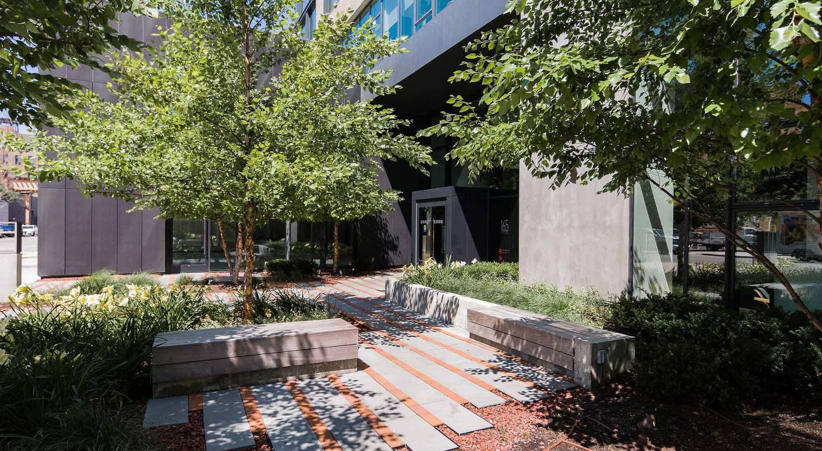 Outdoor courtyard with wooden benches, mature trees providing shade, brick and stone pathways, and modern building facade.