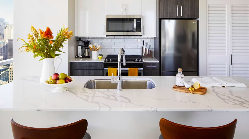 Modern kitchen with white marble island countertop, dual sink, stainless steel appliances, dark cabinetry, and fresh flowers in a white vase.