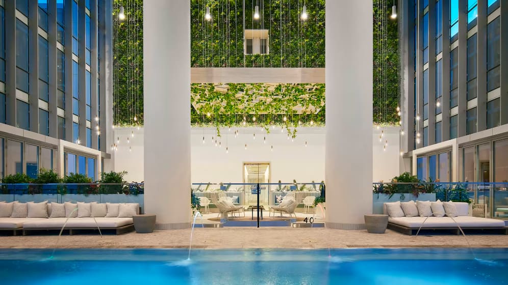 Indoor pool area with blue water, white lounge seating on both sides, tall columns, cascading green plants overhead, and high windows overlooking the city.