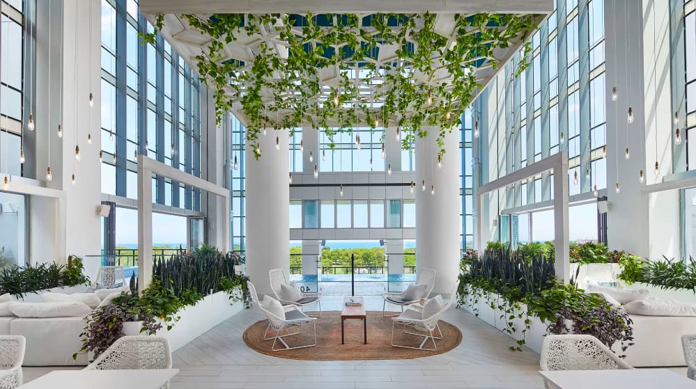 Modern atrium lobby with white columns, hanging plants, large windows, seating areas, and water views in background.
