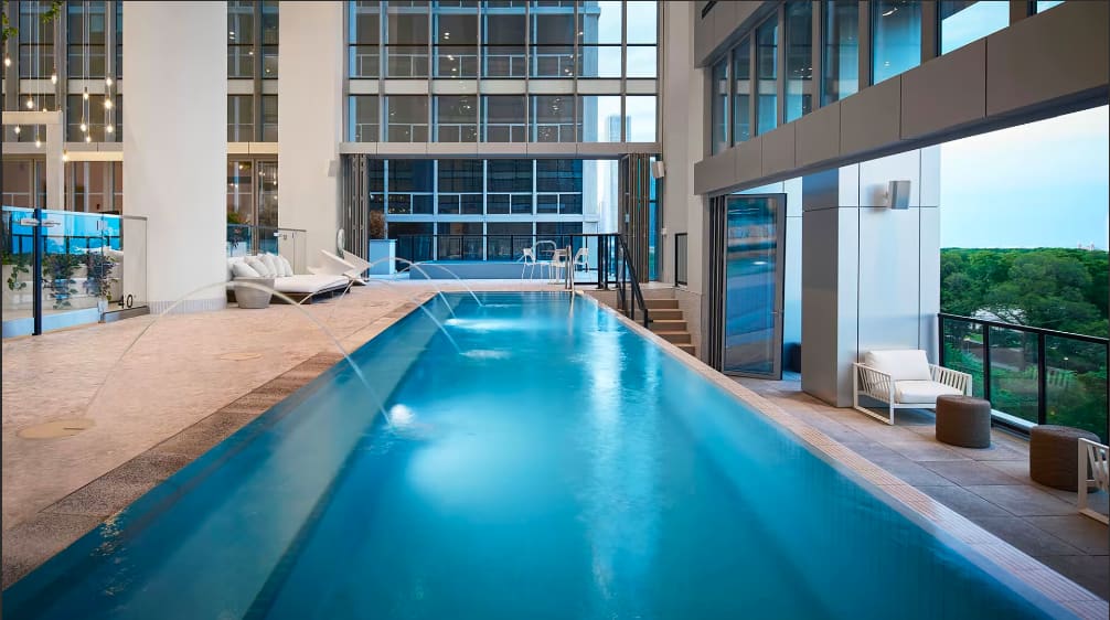 Indoor lap pool with bright blue water, white lounge chairs, floor-to-ceiling windows, and city views in a modern high-rise building.