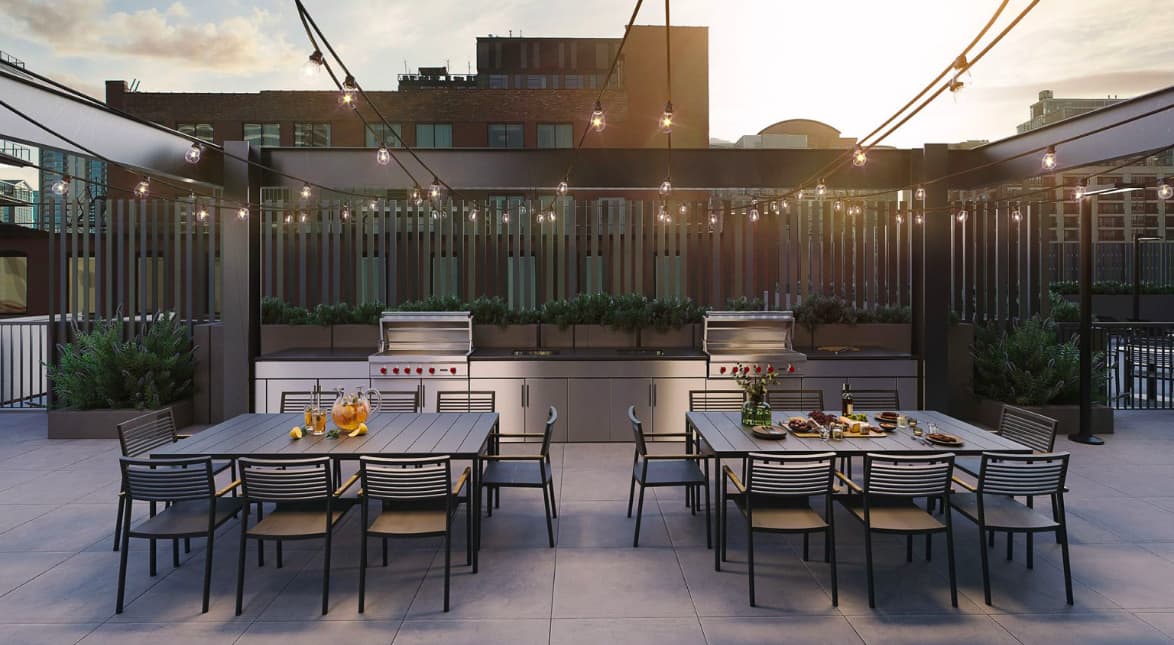 Rooftop terrace with two outdoor kitchen islands, dining tables with black chairs, potted plants, and string lights against an urban backdrop.