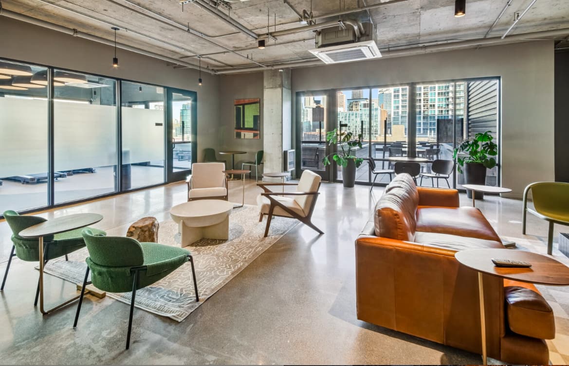 Open-plan lounge with green and orange mid-century chairs, concrete floors, and expansive windows showing downtown Chicago buildings.