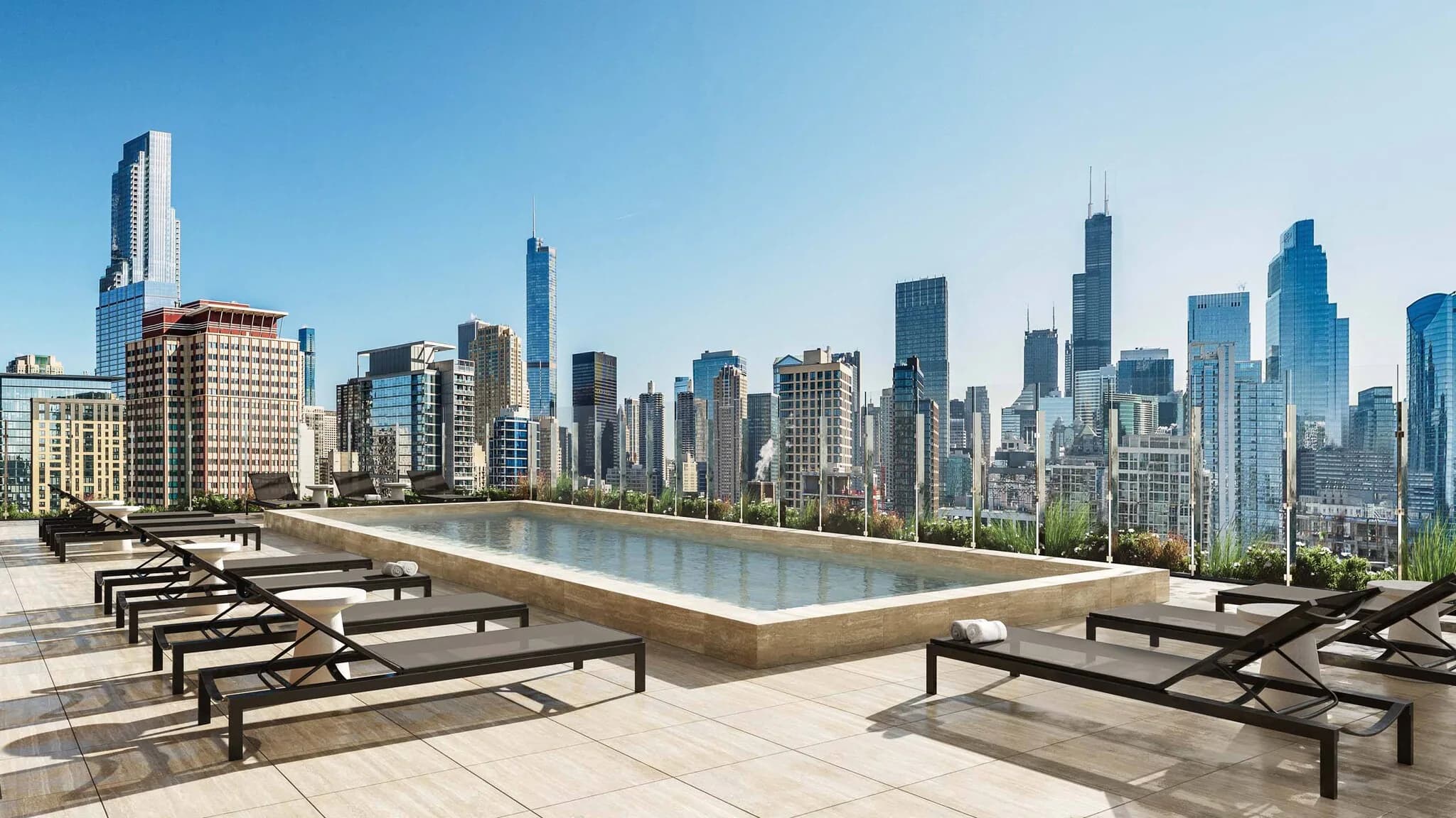 Rooftop pool deck with black lounge chairs, infinity pool, and Chicago skyline backdrop under clear blue sky.