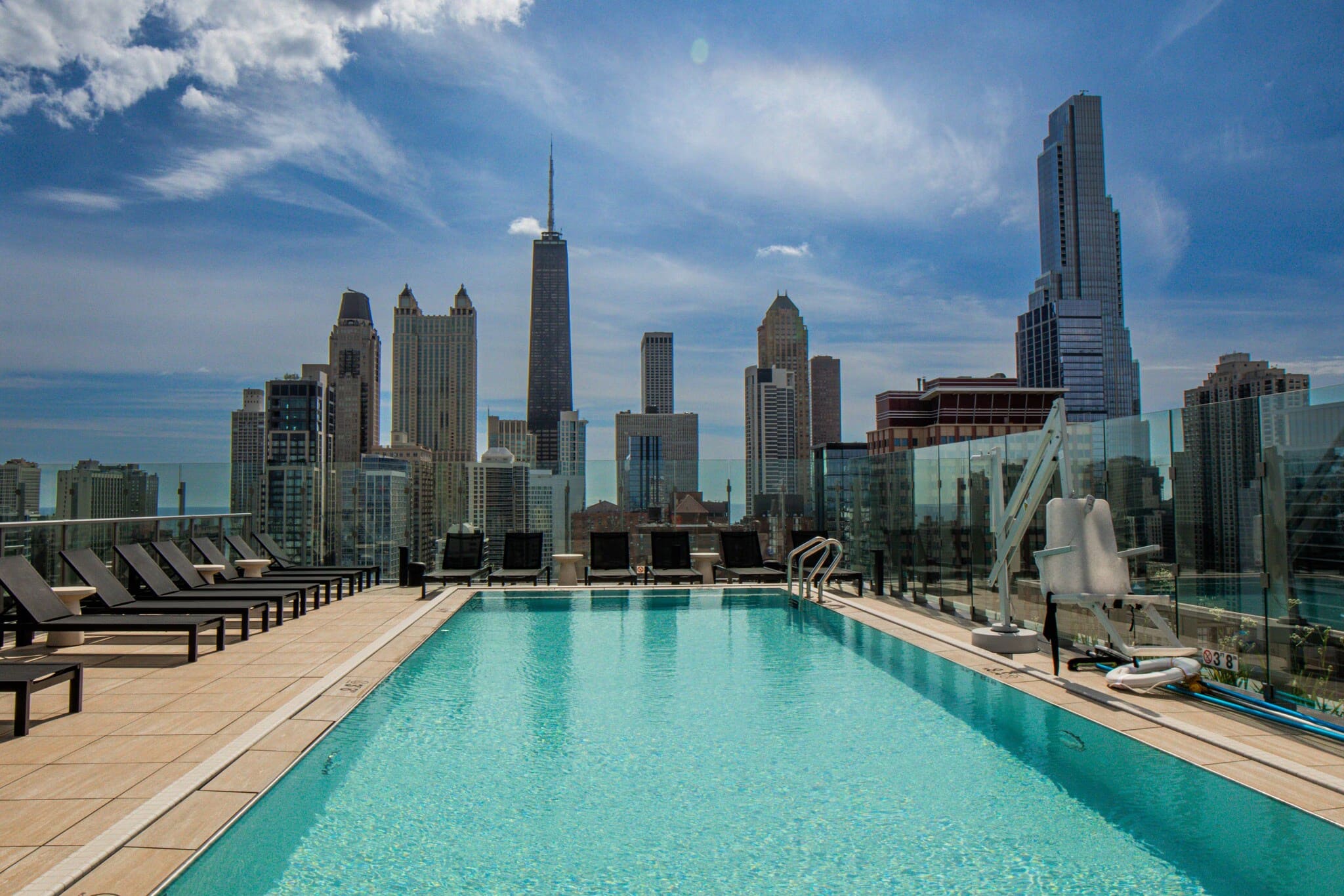 Rooftop swimming pool with turquoise water, lounge chairs, glass railing, and Chicago skyline backdrop under blue sky.