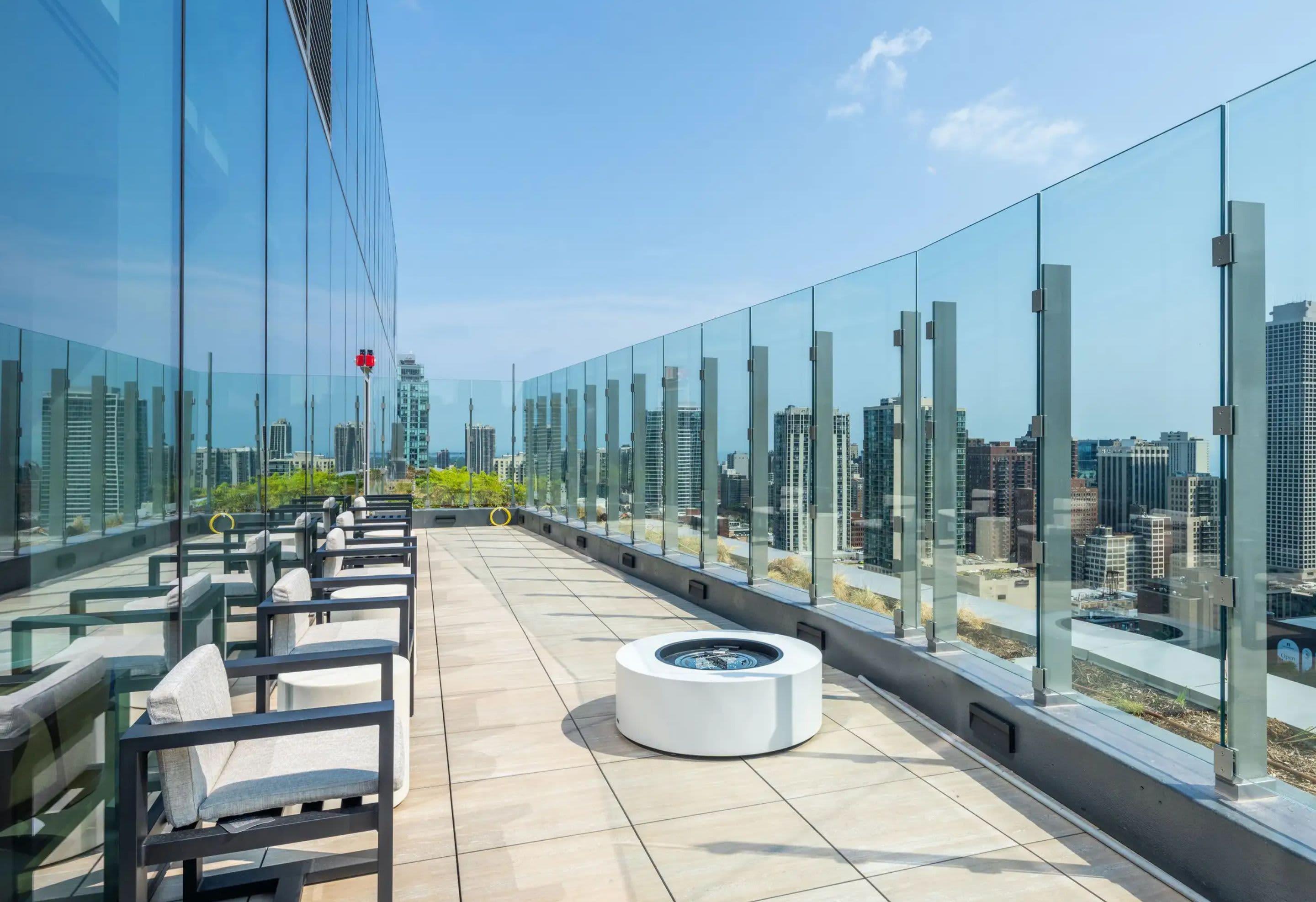 Rooftop terrace with lounge chairs, round white fire pit, and floor-to-ceiling glass railings overlooking Chicago skyline.