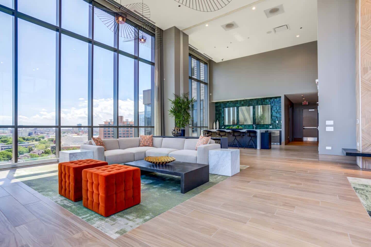 High-rise living room with gray sectional sofa, orange ottomans, black coffee table, floor-to-ceiling windows overlooking cityscape, and wood flooring.