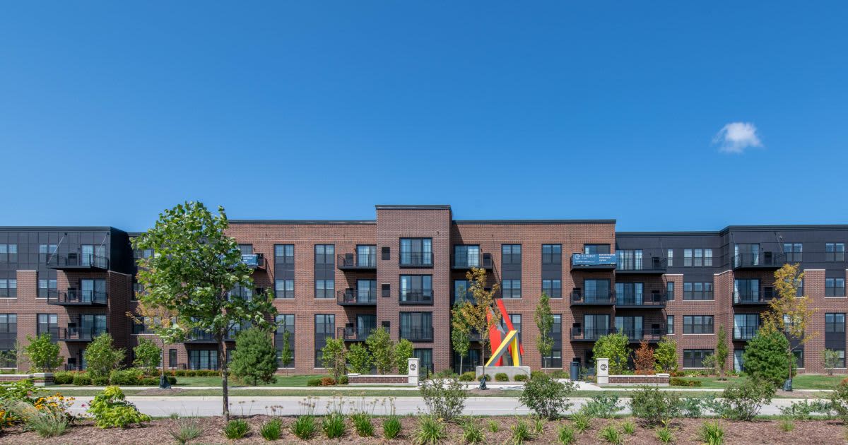Multi-story brick apartment complex with balconies, mature trees, and manicured landscaping under clear blue sky.